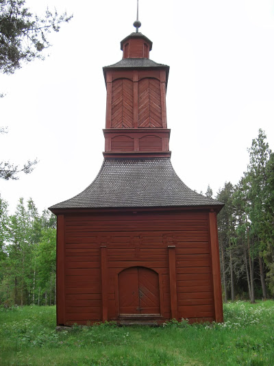 Punaiseksi maalattu puinen kellotapuli. Ympärillä niittyä ja taustalla metsää.